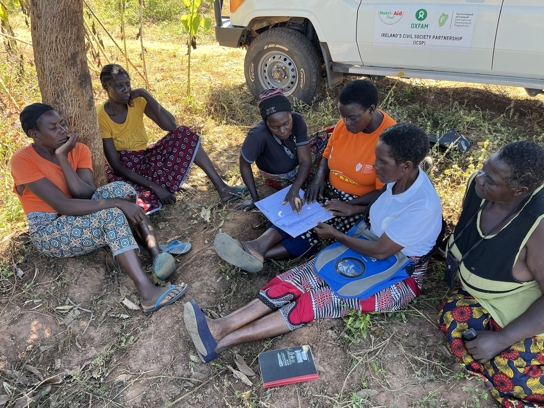 Women farmers reviewing records beside a project vehicle carrying the Nutri-Aid Trust, Oxfam and Irish Aid partnership logos